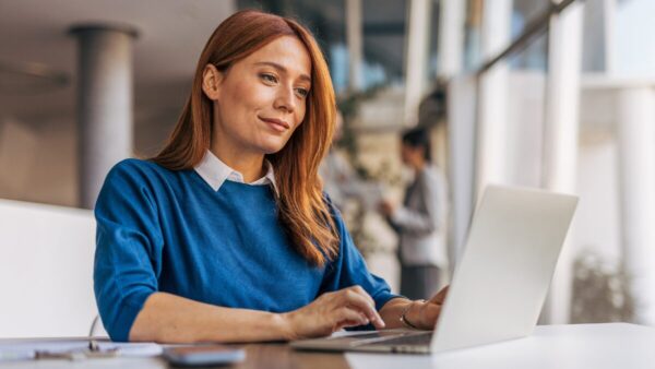 Younge Caucasian woman with auburn hair smiles at laptop in bright airy office environment